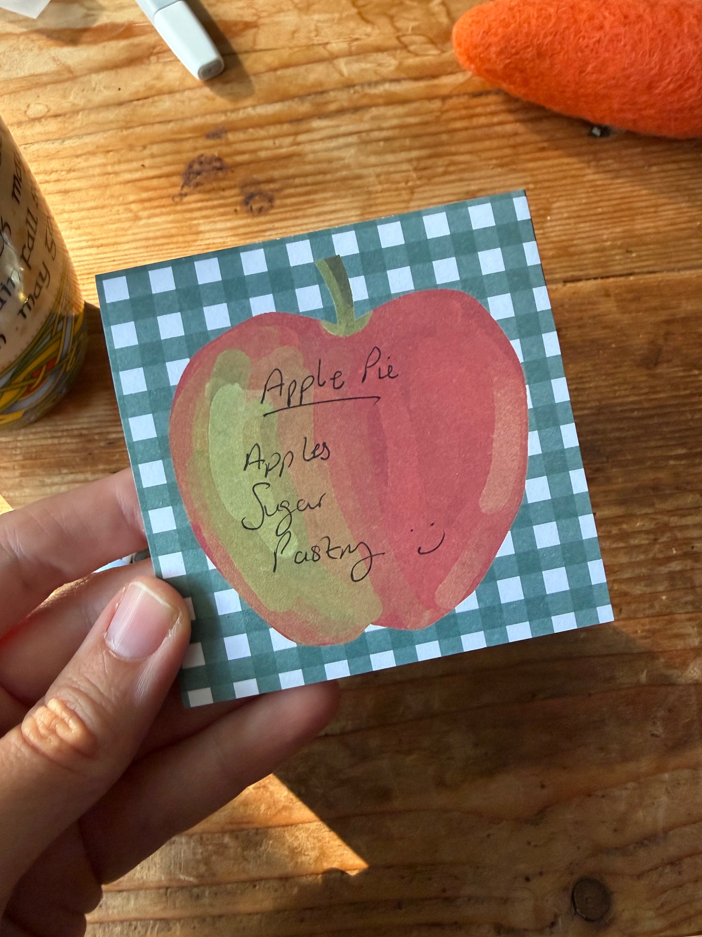A hand holds a square sticky note with a green and white gingham border and a watercolor-style red apple illustration labeled 'Apple Pie' with handwritten notes. The background is a wooden tabletop with other blurred objects around it.